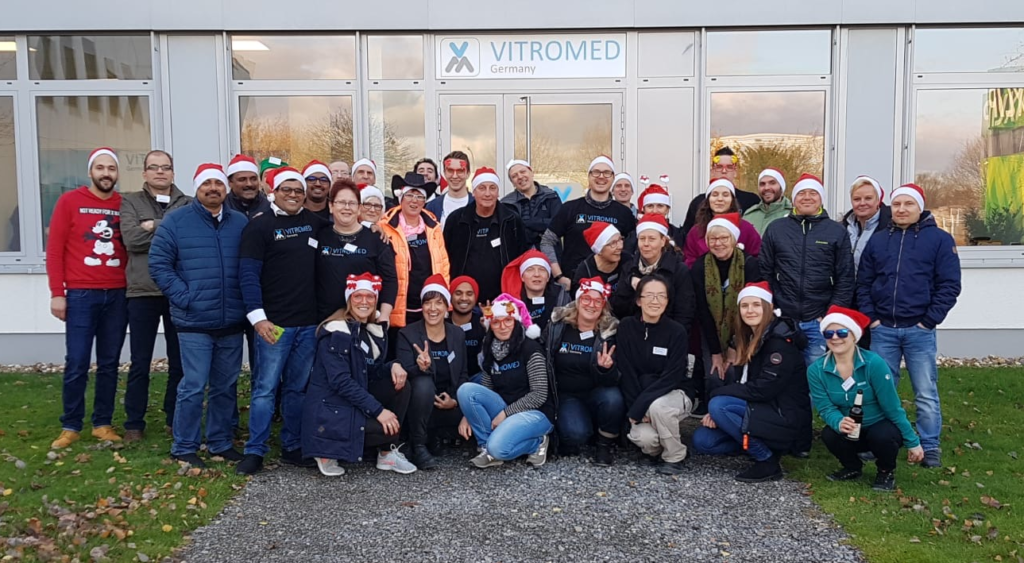 Gruppenfoto vor der Firma VITROMED. Die Personen tragen Weihnachtsmützen und sind gut gelaunt.